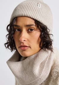 Berretto beige lavorato a maglia con una texture a coste, abbinato a un caldo collo coordinato a coste. Entrambi presentano un'accattivante etichetta minimalista.