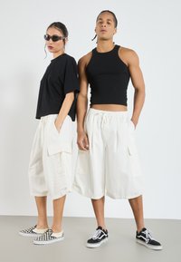 Black cropped t-shirt, cream cargo shorts with side pockets, black tank top, and checkerboard slip-on shoes on the left, black sneakers on the right.