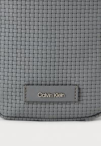 Γκρίζο υφασμάτινο υλικό με υφαντή υφή και ραμμένη ετικέτα που απεικονίζει την ένδειξη "Calvin Klein" σε ασημένια μεταλλικά γράμματα.