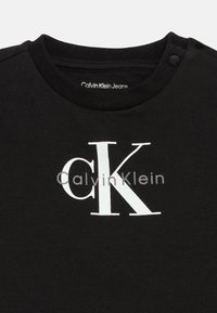 Μαύρη βαμβακερή μπλούζα με λευκό λογότυπο "Calvin Klein" και γραφικό "ck". Στρογγυλή λαιμόκοψη με λεπτομέρεια κουμπιού στον ώμο.
