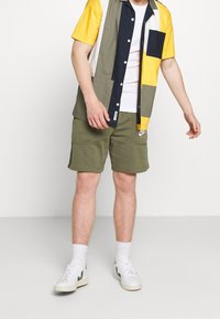 Muž v tričku s krátkým rukávem v barevném blokování, bílém tričku, olivově zelených šortkách s kapsami, bílých ponožkách a bílých teniskách stojí na bílém pozadí.