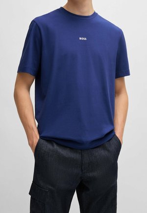 Granatowy bawełniany t-shirt z okrągłym dekoltem i krótkimi rękawami, z małym białym logo "BOSS" na przodzie. Ręce w kieszeniach.