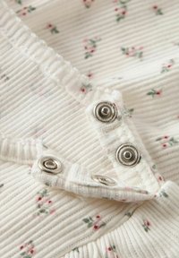 Tissu blanc à côtes avec de petits motifs floraux roses ; présente des boutons-pression argentés et un bord de col volanté. Texture douce.