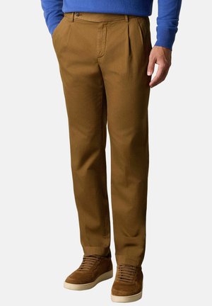 Man die bruine casual broek, bruine suède sneakers en een blauw trui met lange mouwen draagt, met zijn handen in de zakken, staand tegen een effen achtergrond.