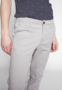 Pantalon gris clair en tissu lisse, coupe ajustée, fermeture par bouton, avec poches latérales. Associé à une chemise foncée à motifs.