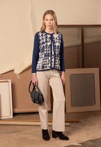 Námornícka modrá a slonovinová kardigán s vzorom psie zuby, v kombinácii s krémovými nohavicami a tmavohnedými čižmami na opätku, držiac malú sivú tašku.