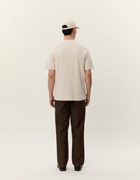 Beige rövidujjú ing, laza fazonban, barna bővebb szárú nadrággal. Könnyű szövettextúra, minimális dizájn, személy bézs sapkát visel.