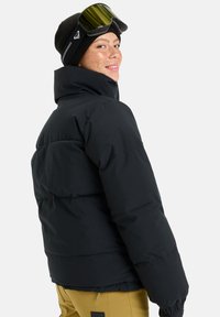 Padded black jacket with a high collar, featuring a textured finish. Worn with a black beanie and ski goggles, highlighting the sporty design.