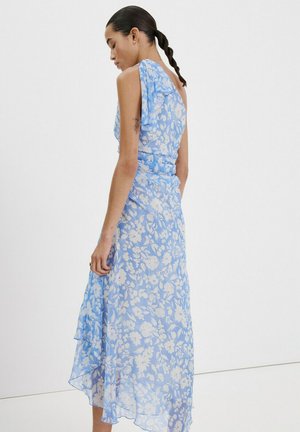 Femme aux cheveux tressés portant une robe midi sans manches bleue et blanche à motif floral avec des détails à volants, se tenant de profil devant un fond uni.