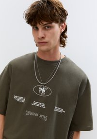 T-shirt in cotone verde oliva con maniche corte, caratterizzato da un design grafico e testo bianco. Catene d'argento sovrapposte aggiungono dettagli.