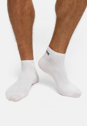 Jambes masculines portant des socquettes blanches avec un texte noir sur le revers, debout sur une surface blanche.