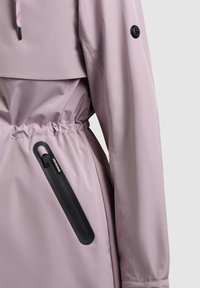 Svetlofialová nepremokavá bunda s elastickým pásom a zvislým čiernym nepremokavým zipsovým vreckom na boku.