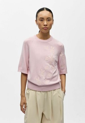 Femme portant un pull à manches courtes rose clair avec des fleurs brodées et un pantalon beige plissé, debout devant un fond blanc uni.