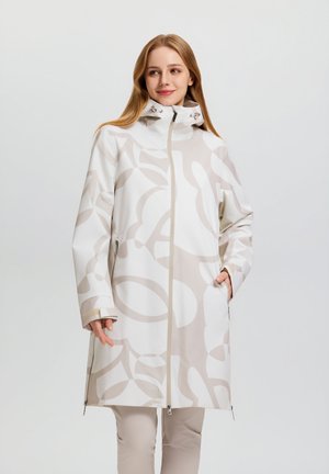 Junge Frau mit langem weißem Mantel mit beigen abstrakten Mustern und hohem Kragen, die mit einer Hand in der Tasche vor einfarbigem Hintergrund steht.
