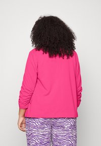 Giacca fucsia con maniche lunghe e dettagli arricciati sulle braccia, abbinata a pantaloni con stampa zebra viola e bianca.