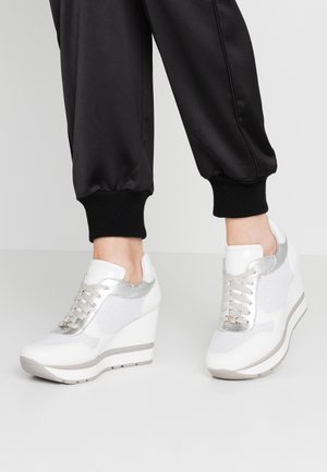 Feet wearing white wedge sneakers with silver accents paired with black cuffed pants against a plain background.