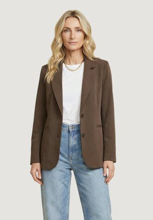 Donna bionda che indossa un blazer marrone sopra una camicia bianca, con jeans azzurri e una collana d'oro, davanti a uno sfondo semplice.