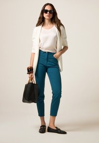 Witte blazer over een lichte top, gecombineerd met smaragdgroene fitted broek. Accessoires zijn zwarte zonnebril, een gestructureerde zwarte tas en gestapelde armbanden.