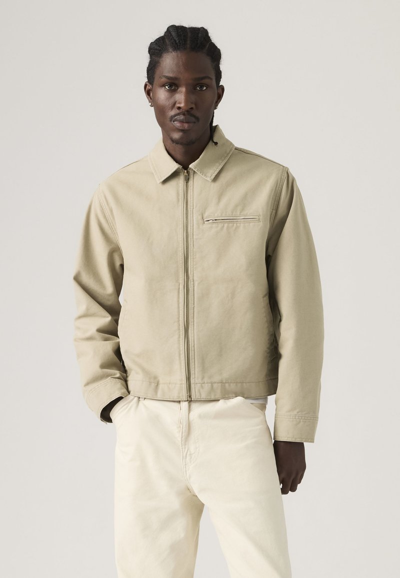 Schwarzer Mann mit geflochtenem Haar trägt eine hellbeige Jacke mit Reißverschluss und cremefarbene Hose, steht mit einer Hand in der Tasche vor einfarbigem Hintergrund.