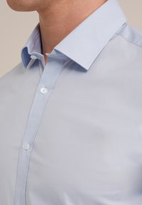 Chemise bleu clair à col étroit, texture lisse, et patte de boutonnage avant avec des boutons blancs. Le tissu semble léger et frais.