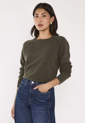 Femme portant un pull vert foncé à manches longues et un jean bleu, debout avec une main dans sa poche devant un fond clair uni.