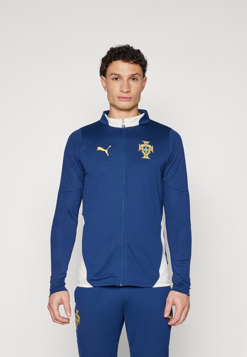 Giacca atletica blu con zip, dotata di pannelli laterali bianchi, con un logo dorato sul petto e un emblema dorato Puma sul lato sinistro.