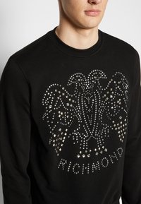 Svart sweatshirt med en silver nitar utsmyckad örn-design och texten "RICHMOND", tillverkad av ett mjukt tyg med en klassisk rundad hals.