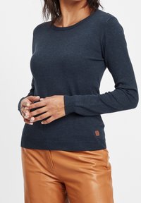 Mörkblå långärmad tröja i ett mjukt tyg med ribbad halsringning och ärmslut; har en liten brun märkespatch vid fållen.