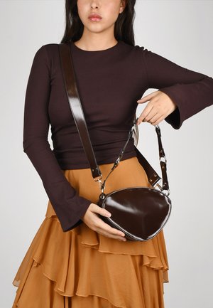 Bolso de mano de cuero marrón con forma curva y acabado brillante. Cuenta con una correa desmontable y clips de metal. Se lleva con una falda marrón con volantes.