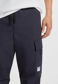 Navy blue cargo pants made of textured fabric, featuring a side pocket and black drawstring. Includes a branded label on the leg.