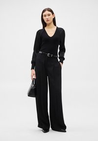 Sieviete, kas valkā melnu V veida kakla tops ar caurspīdīgām piedurknēm, melnas plata piegriezuma bikses, melnu jostu, melnus smailus apavus un tur melnu rokassomu.