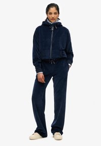 Granatowy dres velurowy z zamkiem błyskawicznym i kapturem z tasiemką, zestawiony z pasującymi spodniami o szerokim kroju. Charakteryzuje się miękką teksturą i luźnym fasonem.
