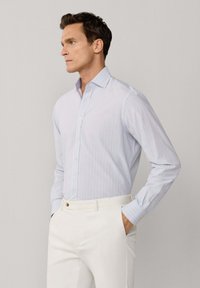 Mann trägt hellblau gestreiftes Hemd mit Knöpfen, in weiße Hose gesteckt, steht mit den Händen in den Taschen und schaut nach links vor einfachem Hintergrund.