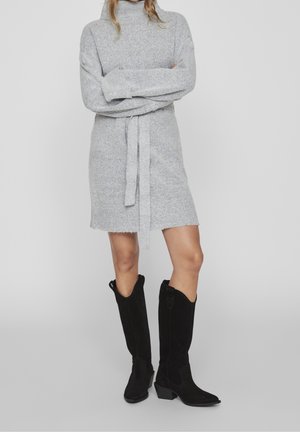 Robe tricotée gris clair avec col roulé et ceinture assortie, présentant une texture douce et un ourlet effiloché, portée avec des bottes en suède noires jusqu'au genou.