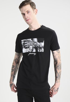 T-shirt en coton noir avec un graphisme central de mains tatouées et chaînes, accompagné du mot "prie" en script blanc.