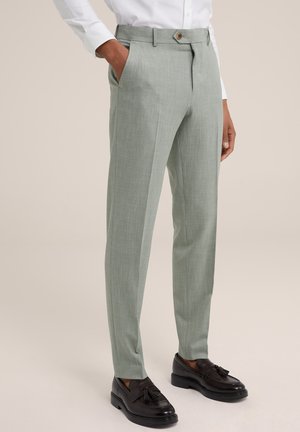 Pantaloni verde chiaro con finitura testurizzata, dotati di una parte anteriore piatta, tasche laterali e chiusura a bottone. Indossati con mocassini neri.