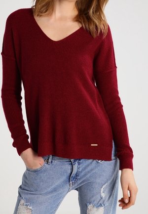 Femme portant un pull bordeaux côtelé à col en V manches longues et un jean bleu clair déchiré, les mains partiellement dans les poches.