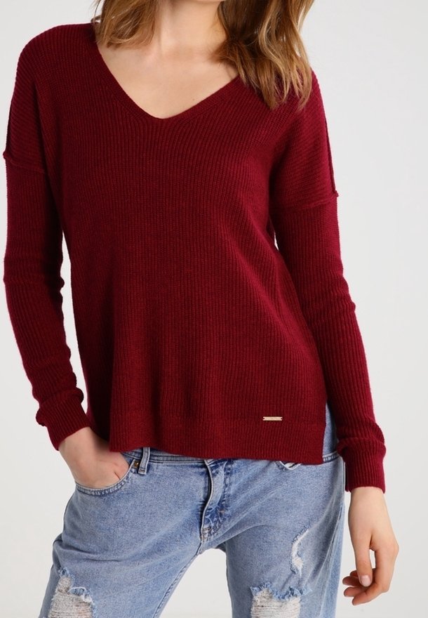 Femme portant un pull bordeaux côtelé à col en V manches longues et un jean bleu clair déchiré, les mains partiellement dans les poches.