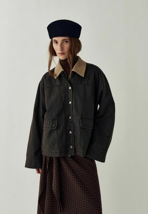Femme portant un béret noir, une veste sombre boutonnée avec un col camel, un pull à col roulé marron et une jupe marron à carreaux, debout devant un fond uni.