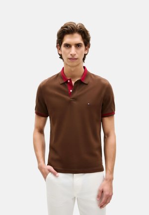 Joven con polo marrón con cuello y puños rojos, de pie con una mano en el bolsillo, vistiendo pantalones blancos sobre un fondo liso.