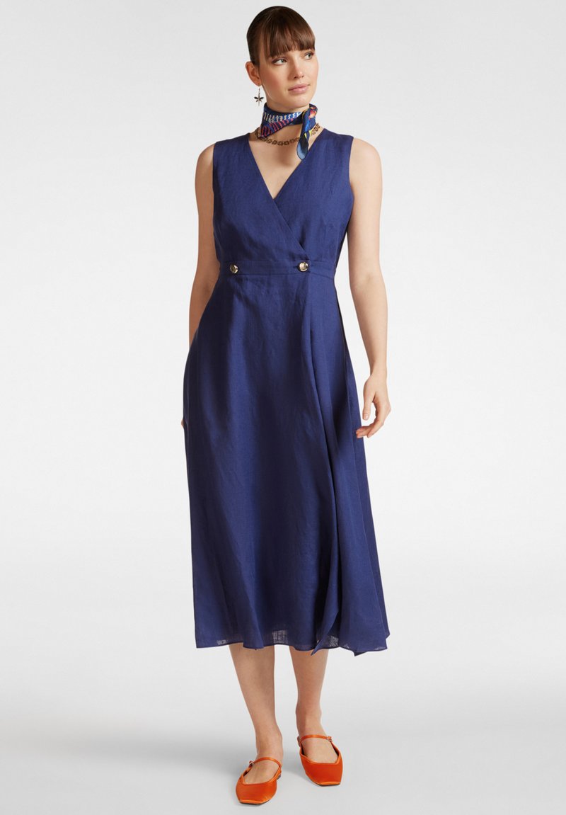 Vestito avvolgente in lino blu navy con scollatura a V, due bottoni dorati in vita e una gonna fluente. Abbinato a delle ballerine arancioni e a una sciarpa colorata.