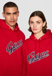Røde hettegensere laget av bomullsblanding, med snor i hetten og en stor hvit og blå "Gant"-logo over brystet.