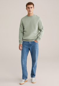 Sudadera verde claro con cuello redondo, combinada con jeans rectos azules y zapatillas blancas con detalles marrones, sobre un fondo neutro.