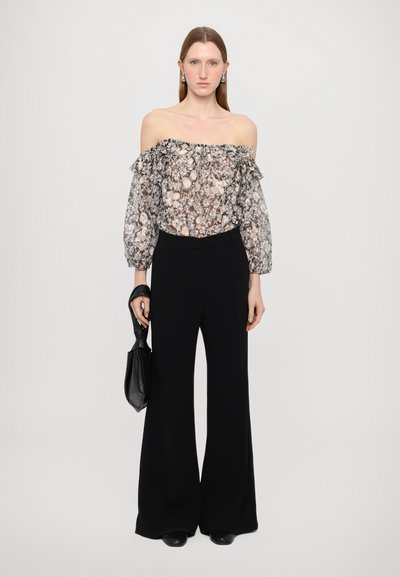 Bluză cu umerii la vedere dintr-un material floral transparent, cu volane, asociată cu pantaloni negri cu talie înaltă și croi larg. Poartă o geantă neagră în mână.