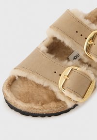 Bež sandaalit kahekordsete rihmadega, pehme karvase voodriga, kuldsete pandlatega ja musta kummitaldaga. Omab siledat sametist materjali ja ümarate nurkadega disaini.