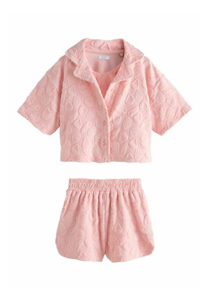 Fiatal lány rózsaszín texturált ruhában mezítláb sétál a homokos tengerparton, háttérben óceán és sziklák, tiszta ég alatt.