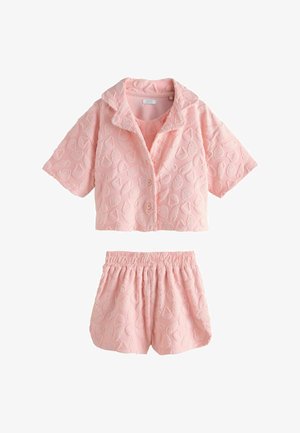 Junges Mädchen in pinkfarbenem, strukturiertem Outfit läuft barfuß am Sandstrand mit Ozean und Felsen im Hintergrund unter klarem Himmel.