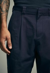 Marineblauwe broek van gestructureerde stof, met een platte voorkant en riemlussen. Een hand ligt op het bovenbeen, waardoor de pasvorm te zien is.