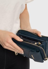 Portafoglio in pelle blu navy con finitura testurizzata, zip dorate e più scomparti. Tenuto in una mano, parzialmente aperto per rivelare l'interno.