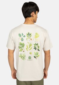Naturliches Baumwoll-T-Shirt in Off-White, mit detaillierten grünen botanischen Illustrationen auf der Rückseite und einem kleinen Logo am unteren Rand.